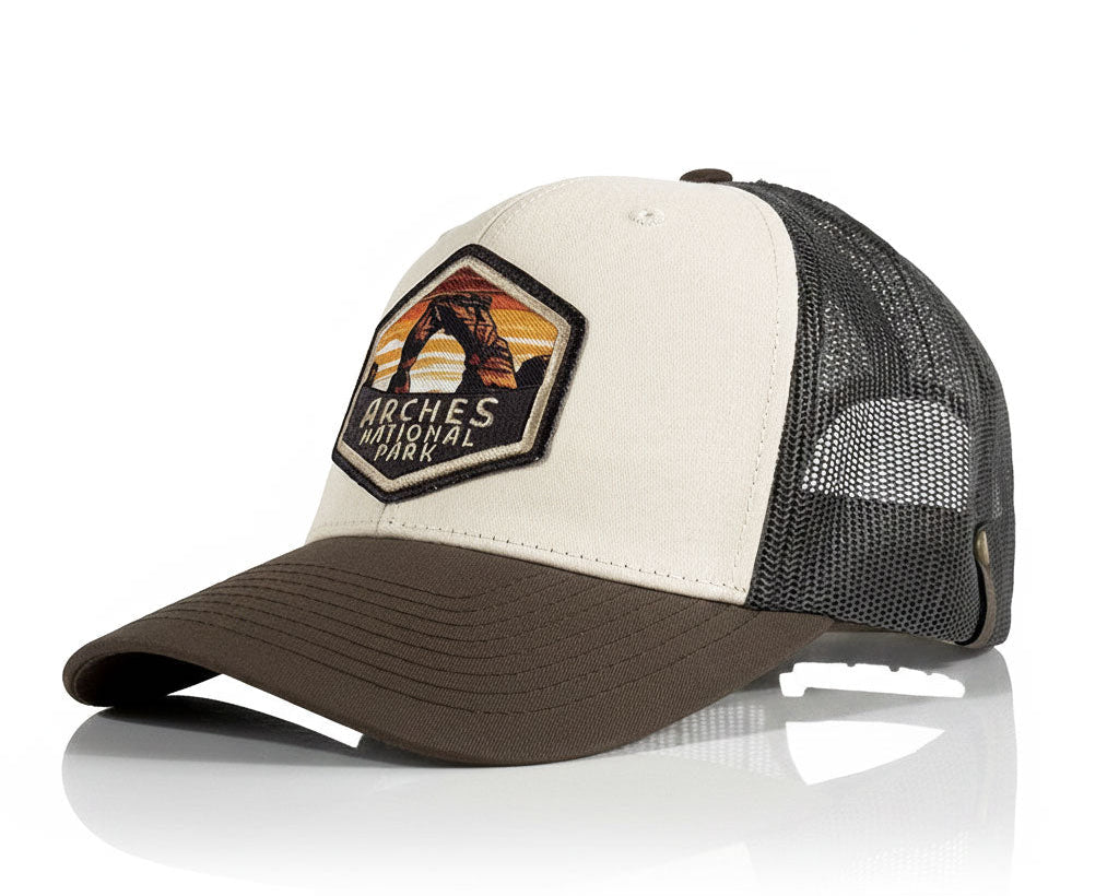 A trucker-style baseball cap with a cream front panel, dark brown brim, and black mesh back. The front panel features a hexagonal patch for 'Arches National Park' with a sunset and horse design.