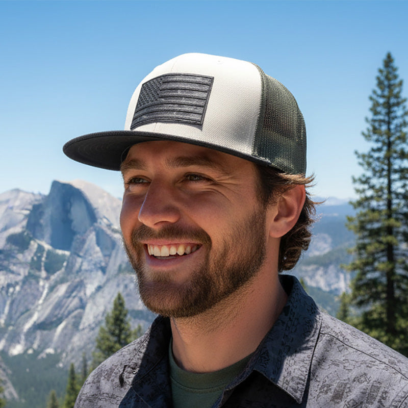 Man wearing a cap with an American flag design, standing in front of mountains and trees.