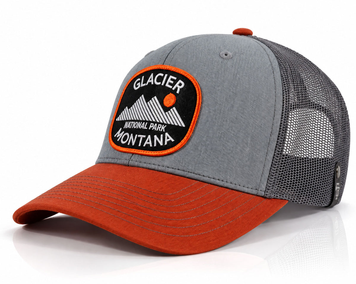 A gray and rust-orange trucker cap with a black mesh back and a circular Glacier National Park Montana patch on the front.