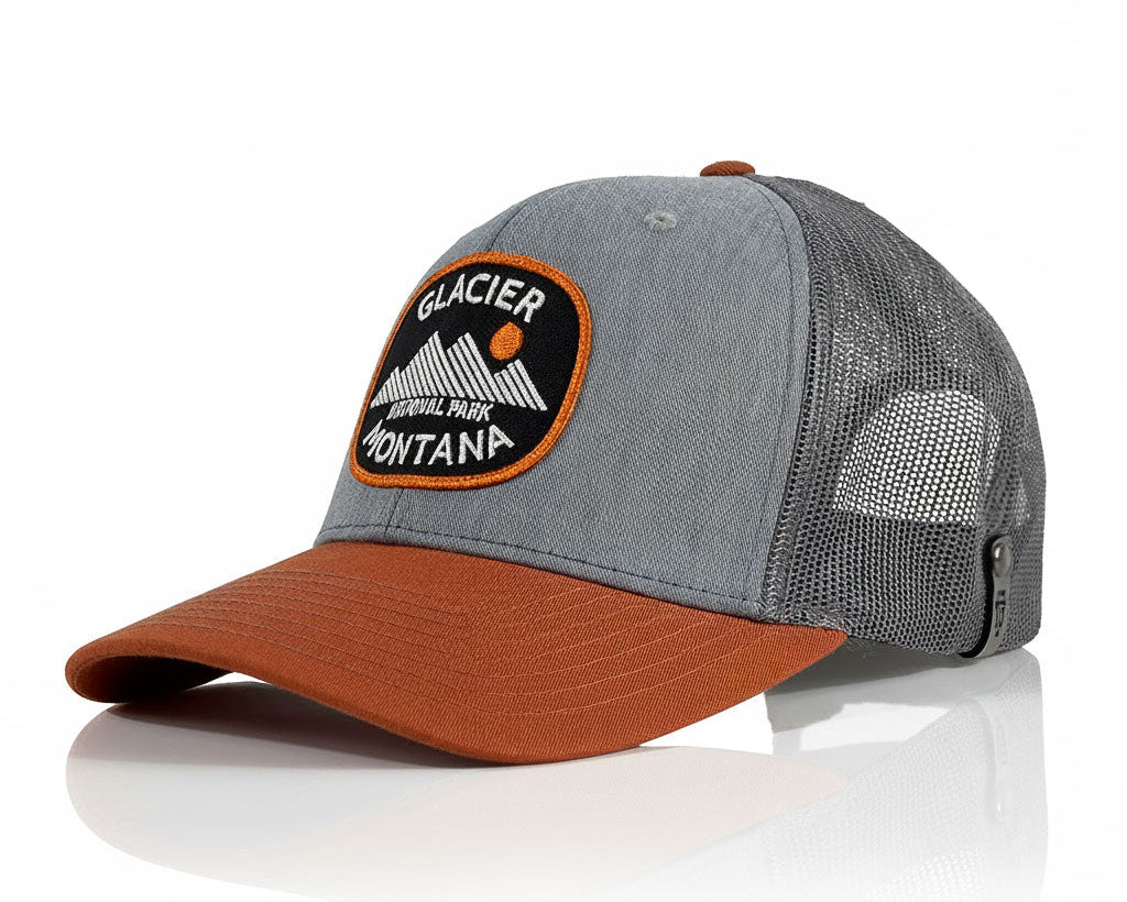 A Glacier National Park Montana trucker hat with a gray twill front panel, dark gray mesh back, and a rust-brown curved brim. The front features an embroidered patch with a black background, white mountain graphic, and orange border.