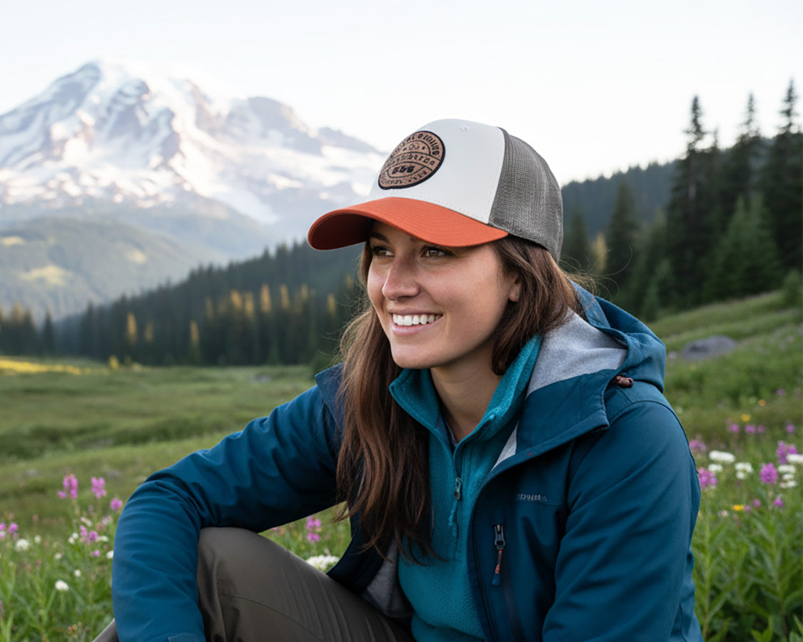 Woman wearing a cap and blue jacket sitting in a field with mountains in the background