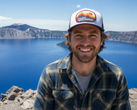 Man wearing a 'Pacific Northwest' cap with a scenic background of a lake and mountains.
