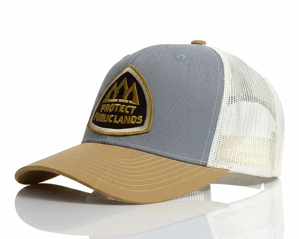 A trucker-style baseball cap with a gray front panel, tan visor, and off-white mesh back. The front panel features a shield-shaped embroidered patch with a mountain graphic and the text 'PROTECT PUBLIC LANDS'.
