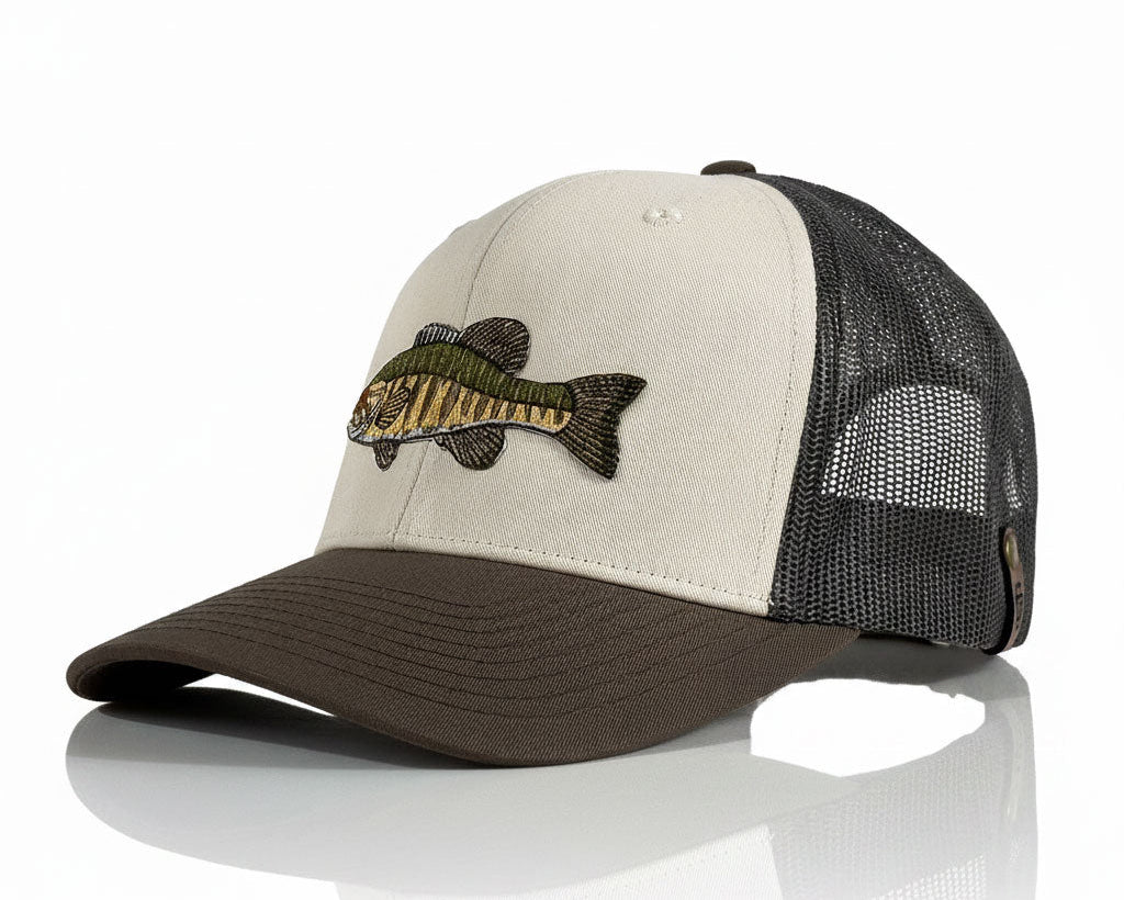 A trucker-style baseball cap with a light beige front panel, dark brown visor, and black mesh back. An embroidered fish patch is stitched on the front.