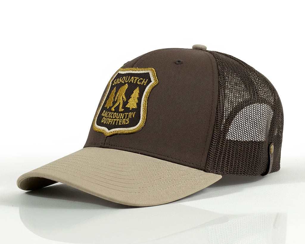 A brown and beige trucker hat with a mesh back, featuring a shield-shaped embroidered patch on the front that reads 'SASQUATCHES & CUCKOUNTRY OUTFITTERS' with a silhouette of a big cat and trees.