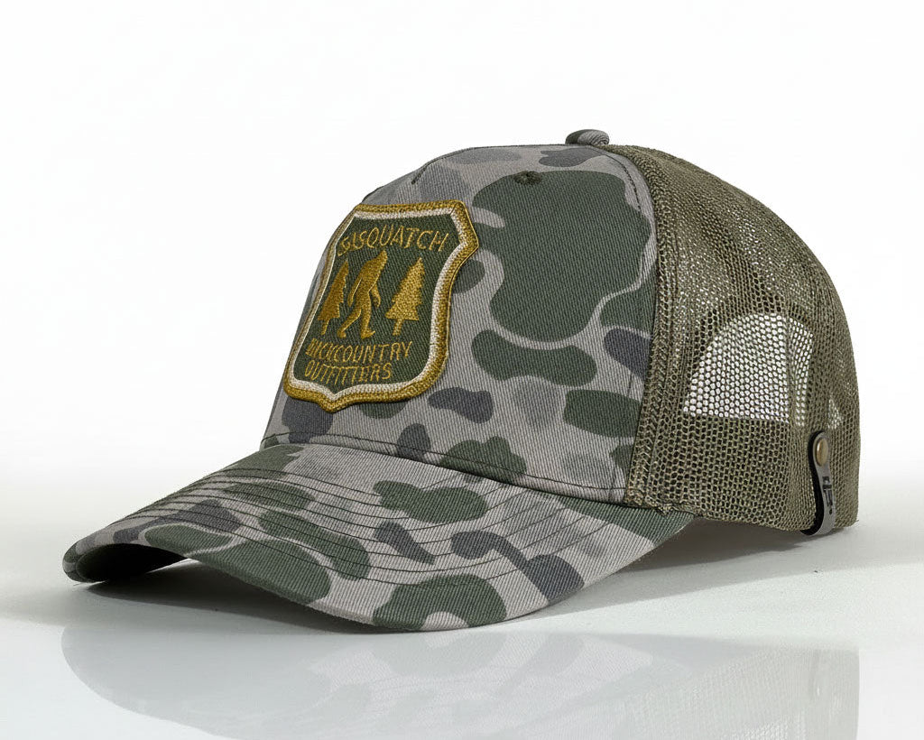 A camouflage trucker hat with a gray and green camo pattern on the front panel and brim, olive green mesh back, and a gold embroidered patch on the front that reads 'CASQUATCH BASKETCOUNTRY OUTFITS'.