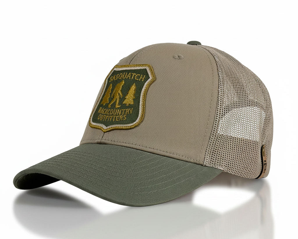 A structured trucker-style baseball cap with a tan front panel, olive green brim, and olive green mesh back. It features a prominent embroidered patch on the front with the text 'CAYSQUATCH BACKCOUNTRY DEFFITORS' and an illustration of three figures hiking up a mountain.