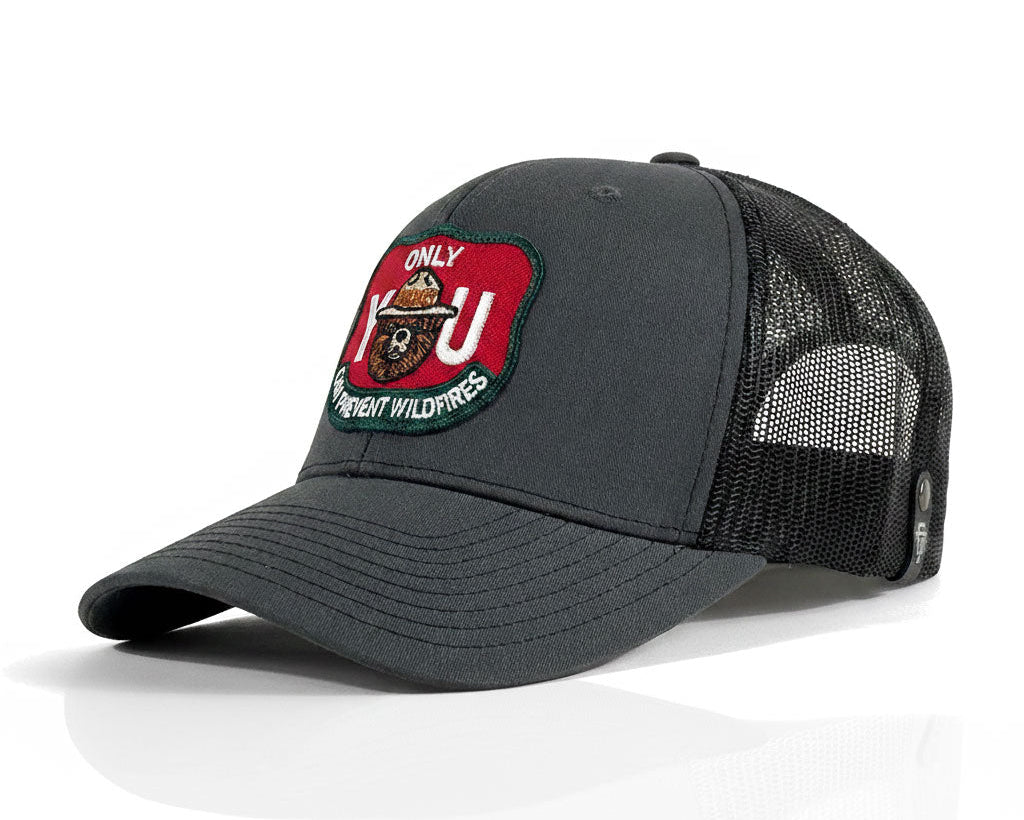 A dark gray trucker hat with a curved brim, black mesh back, and a red, green, and white embroidered patch on the front that reads 'Only You Bring Wildfires'.