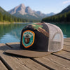Camo mesh trucker hat with Smokey Bear patch on dock by clear lake and mountain view