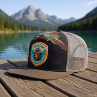 Camo mesh trucker hat with Smokey Bear patch on dock by clear lake and mountain view