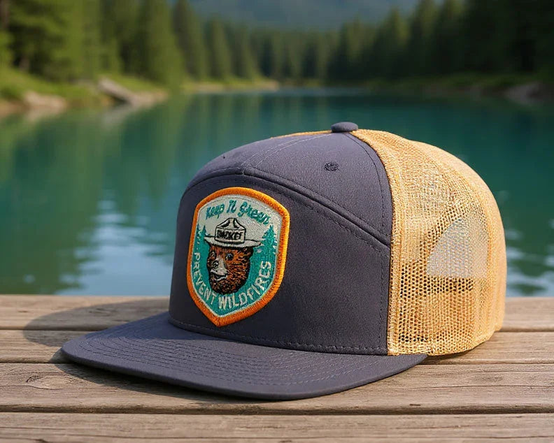 Trucker hat with a patch on a wooden surface in front of a scenic lake and forest.