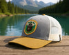 Gray and yellow cap with a patch on a wooden surface with a scenic background of a lake and mountains.