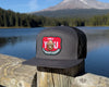 Black cap with a red and green patch on a wooden post by a lake with mountains in the background