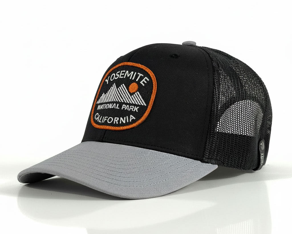 A black and gray trucker hat featuring a circular embroidered Yosemite National Park patch on the front, set against a clean white background with a soft reflection.