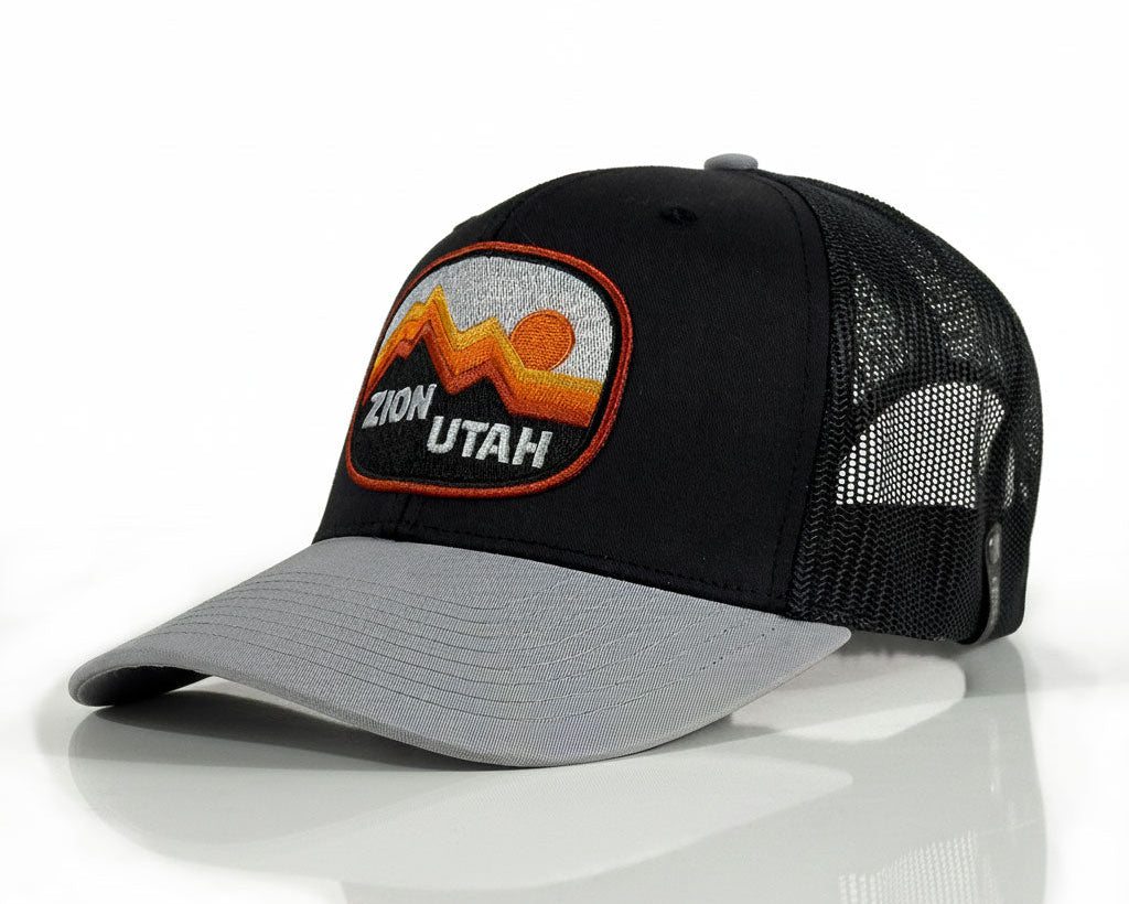 A black and gray trucker hat with a curved gray brim, black mesh back, and a prominent embroidered 'Zion Utah' patch on the front.