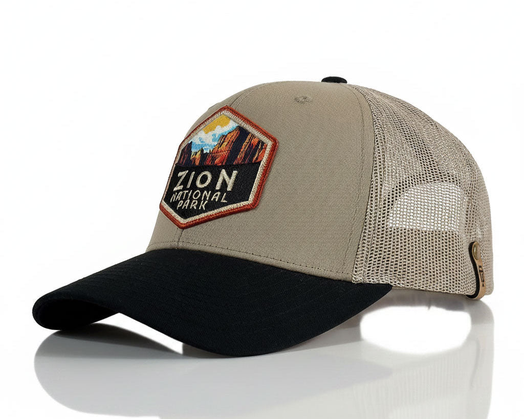 A beige and black trucker hat featuring an embroidered 'Zion National Park' patch on the front, set against a clean white background with a soft reflection.
