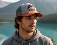 A man wearing a Glacier Mountain brand trucker hat with a gray and orange brim, standing outdoors in front of a scenic mountain and turquoise lake background.