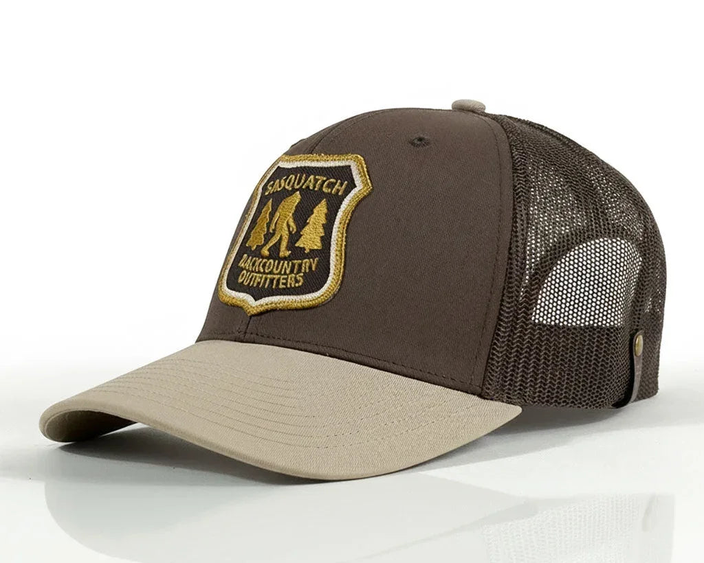 A brown and beige trucker hat with a mesh back, featuring a shield-shaped embroidered patch on the front that reads 'SASQUATCH BACKCOUNTRY OUTFITTERS' in gold and black.