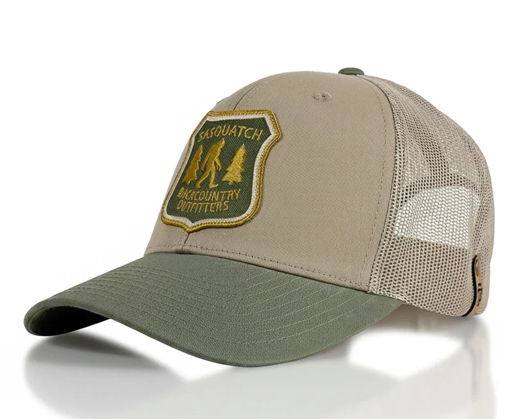 A trucker-style baseball cap with a tan front panel, olive green visor and back mesh, and a green and gold embroidered patch on the front that reads 'SASQUATCH BACKCOUNTRY OUTFITTERS'.