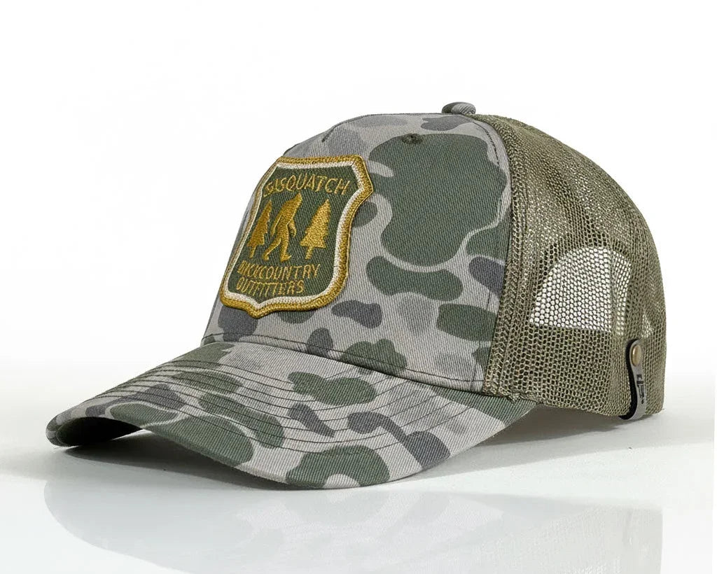 A camouflage trucker hat with green and gray camo print on the front and brim, olive green mesh back panels, and a gold embroidered patch on the front that reads 'SASQUATCH BACKCOUNTRY OUTFITTERS'.