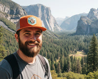 Man with a beard wearing an orange cap with a logo, standing in front of a scenic mountain landscape.