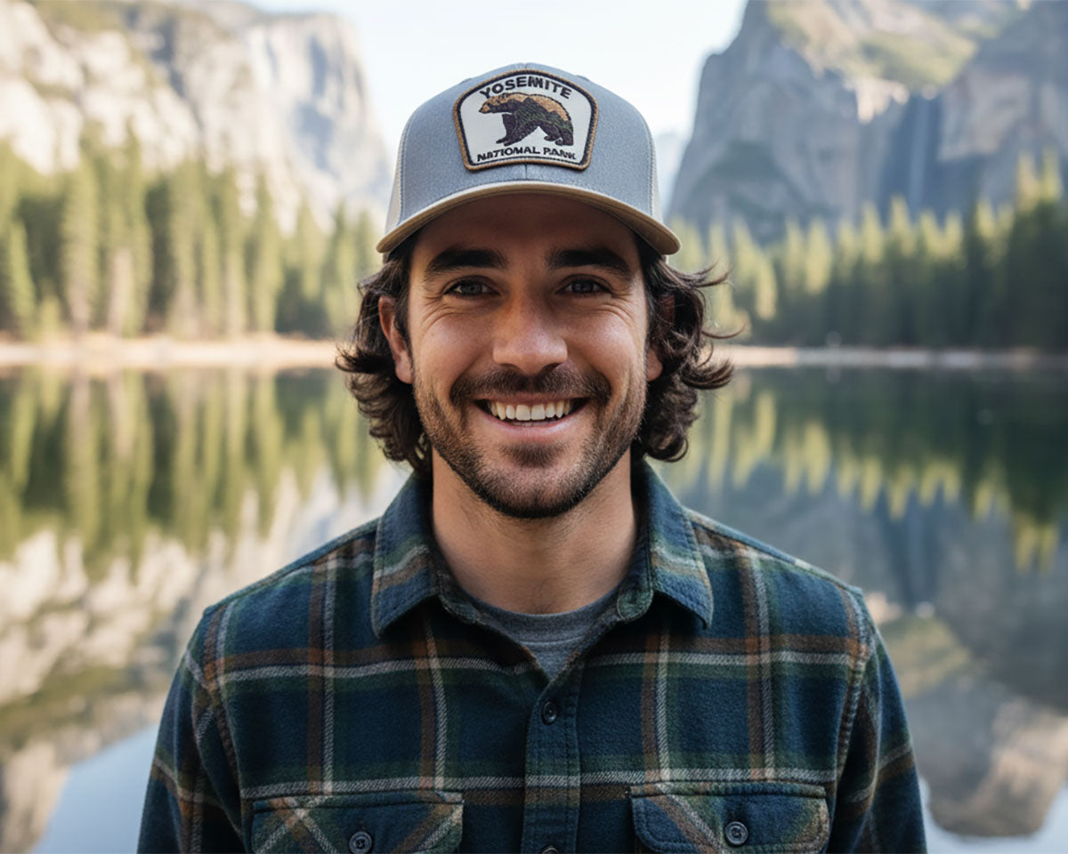 A smiling man wearing a gray Yosemite baseball cap and a green plaid flannel shirt stands in front of a mountain lake background.
